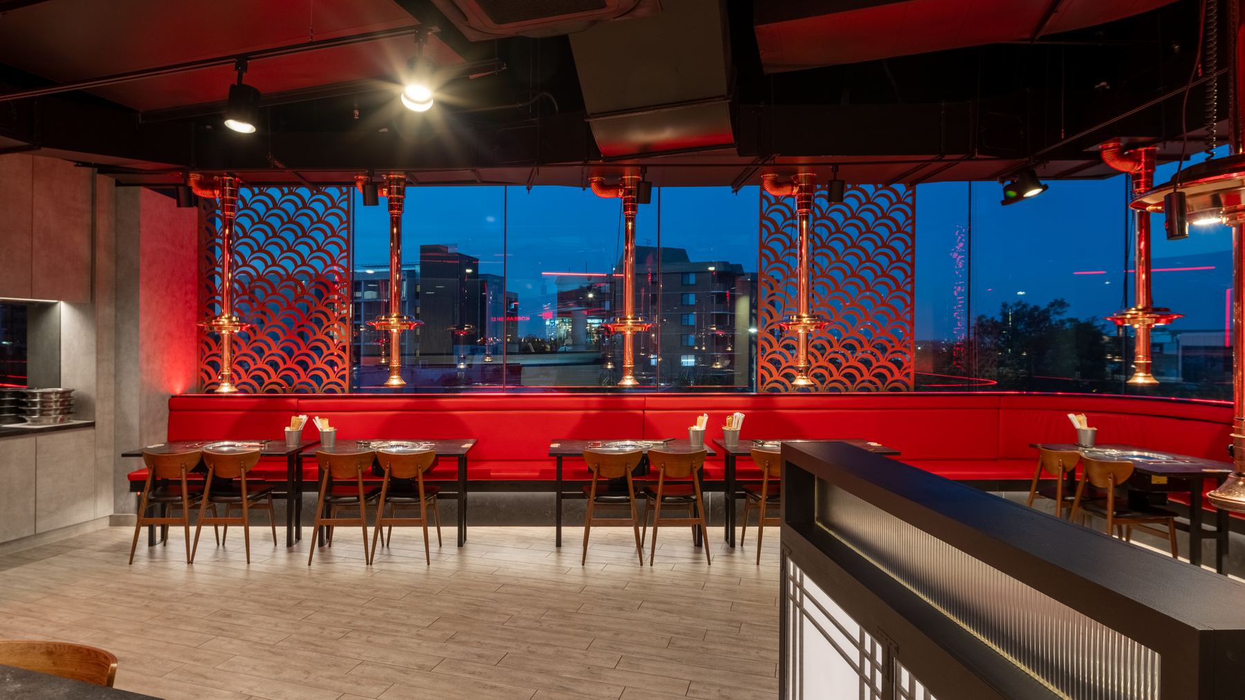 Red banquette booths along full-height glazing at dusk, copper extraction hoods over each table